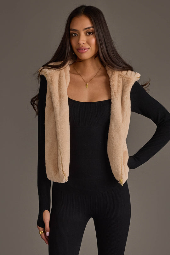A woman with long dark hair wears the Panache Apparel Collection Beige Faux Fur Vest over a fitted black long-sleeve outfit, standing against a plain gray background and smiling slightly.