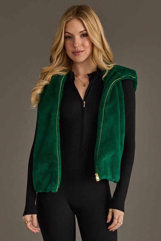 A woman with long blonde hair stands against a plain gray background, wearing the Panache Apparel Collection Green Faux Fur Vest over a black zip-up top and black pants.