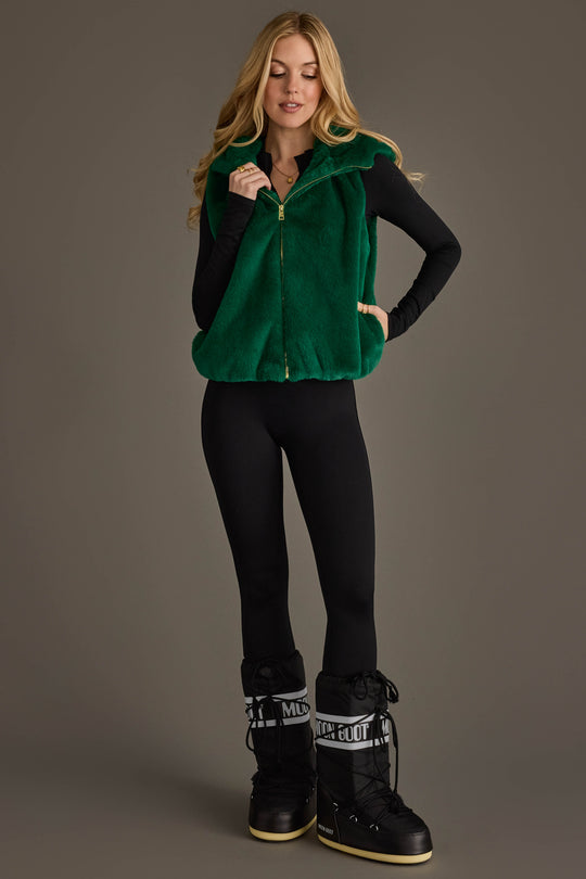 A woman poses against a plain gray background, wearing the Panache Apparel Collection Green Faux Fur Vest layered over a black long-sleeve bodysuit and paired with black snow boots. She has long blonde hair and touches the collar of her vest.