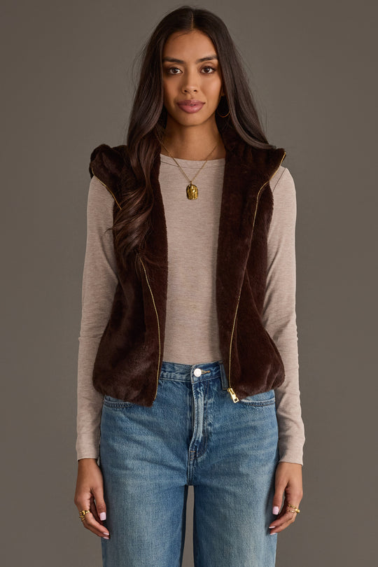 A woman with long brown hair is wearing the Panache Apparel Collection Dark Brown Faux Fur Vest over a beige long-sleeve shirt and blue jeans, standing against a plain gray background.