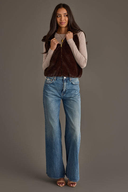 A woman stands against a gray background wearing a long-sleeve beige top, wide-leg blue jeans, brown sandals, and the Panache Apparel Collection Dark Brown Faux Fur Vest, with her long hair down and hands holding her vest.