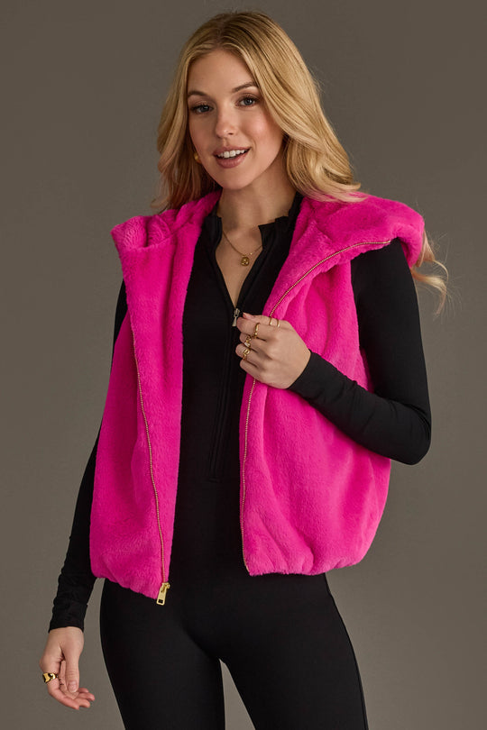 A woman poses and smiles against a neutral background, wearing the Panache Apparel Collection Fuchsia Faux Fur Vest layered over a black long-sleeve outfit.