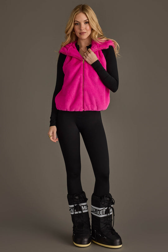 A woman wearing the Panache Apparel Collection Fuchsia Faux Fur Vest over a black long-sleeve top, paired with black leggings and black snow boots, stands against a plain brown background, smiling with her hands near her waist.