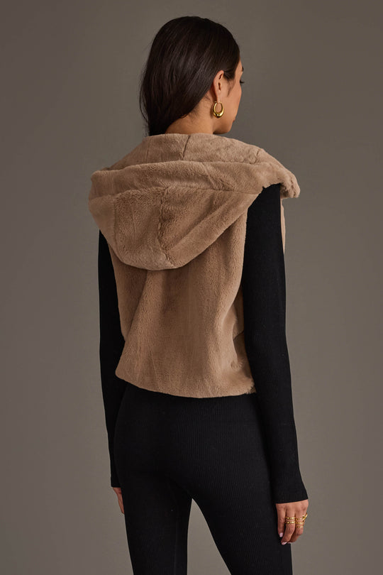 A person with long dark hair is shown from the back against a plain gray background, wearing the Panache Apparel Collection Light Brown Faux Fur Vest, a sleeveless, hooded style, layered over a black long-sleeve outfit.