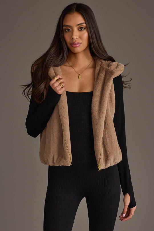 A woman models the Panache Apparel Collection’s Light Brown Faux Fur Vest over a black long-sleeve outfit, standing against a plain gray background and gazing at the camera while holding the collar of the vest with one hand.