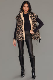 A woman stands against a gray background, modeling the Panache Apparel Collection’s Tan & Brown Leopard Fleece Vest layered over a black long-sleeve top, paired with black leather pants and knee-high black boots. She poses confidently with one hand on her hip, her long hair styled down.