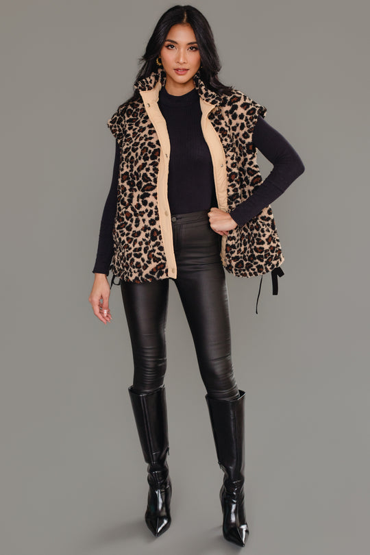 A woman stands against a gray background, modeling the Panache Apparel Collection’s Tan & Brown Leopard Fleece Vest layered over a black long-sleeve top, paired with black leather pants and knee-high black boots. She poses confidently with one hand on her hip, her long hair styled down.