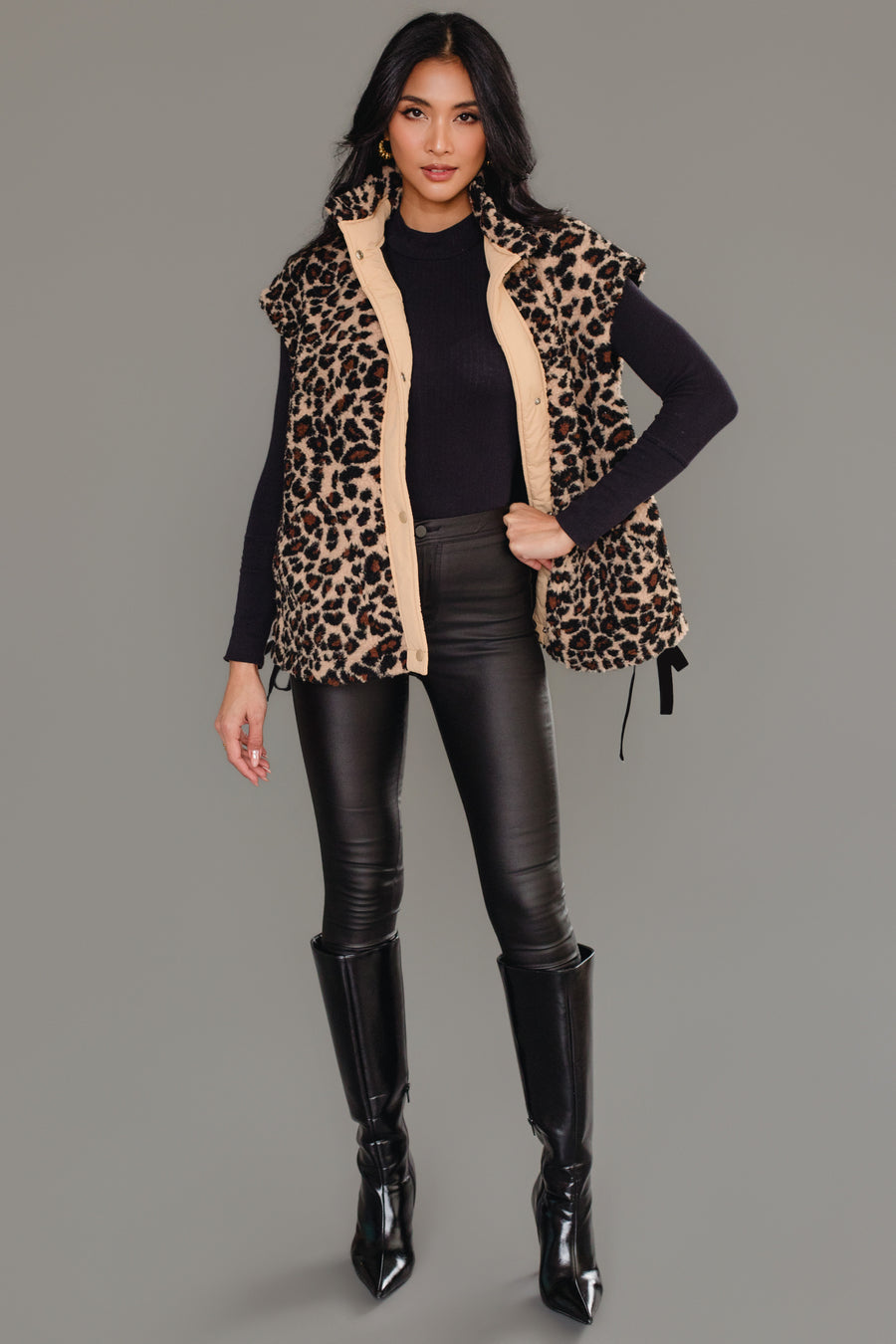 A woman stands against a gray background, modeling the Panache Apparel Collection’s Tan & Brown Leopard Fleece Vest layered over a black long-sleeve top, paired with black leather pants and knee-high black boots. She poses confidently with one hand on her hip, her long hair styled down.