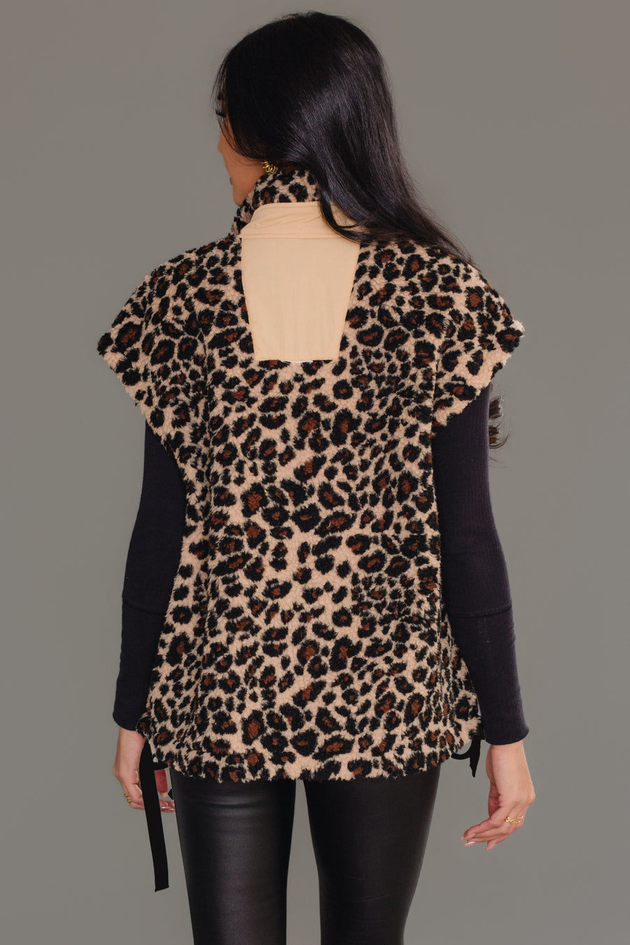 A woman faces away, wearing the Panache Apparel Collection Tan & Brown Leopard Fleece Vest, featuring short sleeves, a high collar, and a rectangular beige patch on the back. The vest is styled over a black long-sleeve top with black pants against a plain gray background.