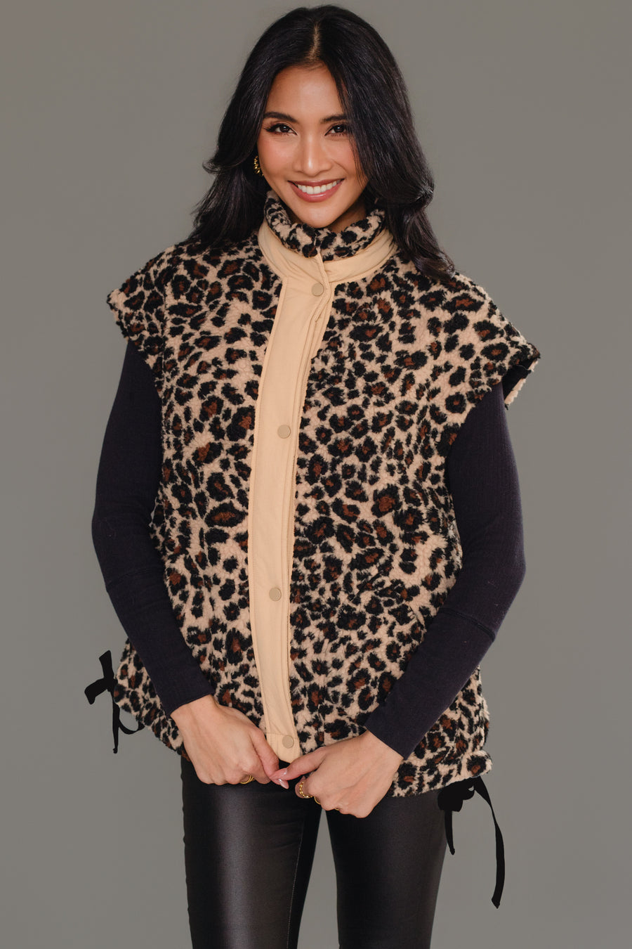A woman smiles, modeling the Panache Apparel Collection’s Tan & Brown Leopard Fleece Vest, which features beige trim. She wears it over a black long-sleeve top and black pants, standing against a plain gray background.