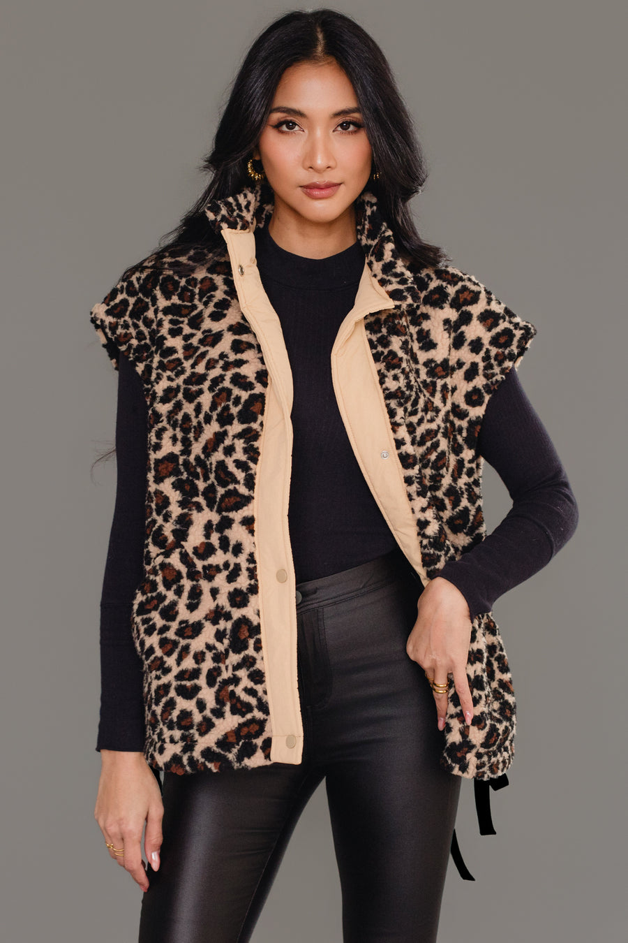 A woman stands confidently against a plain gray background, wearing the Panache Apparel Collection Tan & Brown Leopard Fleece Vest over a black turtleneck sweater and black leather pants.