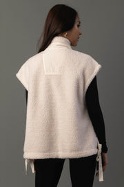 A woman with long brown hair, wearing the Panache Apparel Collection Ivory Fleece Vest over a black long-sleeve shirt and black pants, stands facing away against a plain gray background.