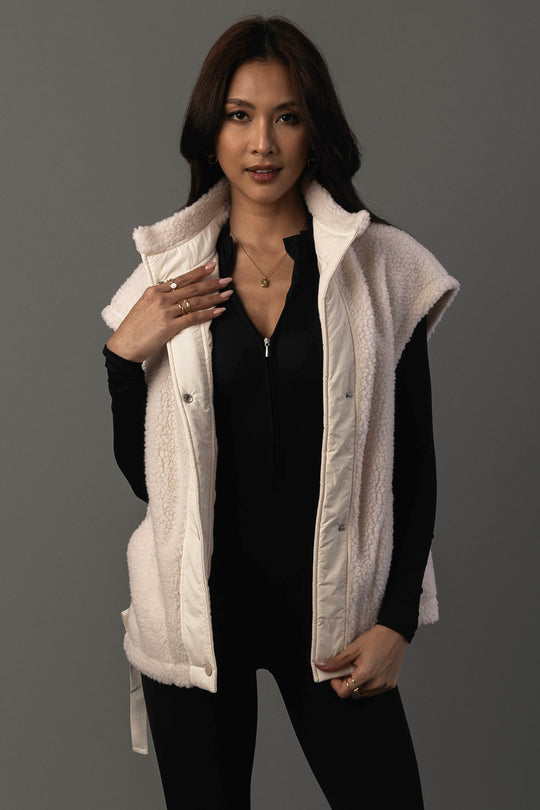 A woman with long dark hair models the Panache Apparel Collection Ivory Fleece Vest, a sleeveless white piece layered over a black outfit. She poses against a plain gray background, touching the vest and accessorizing with gold jewelry.