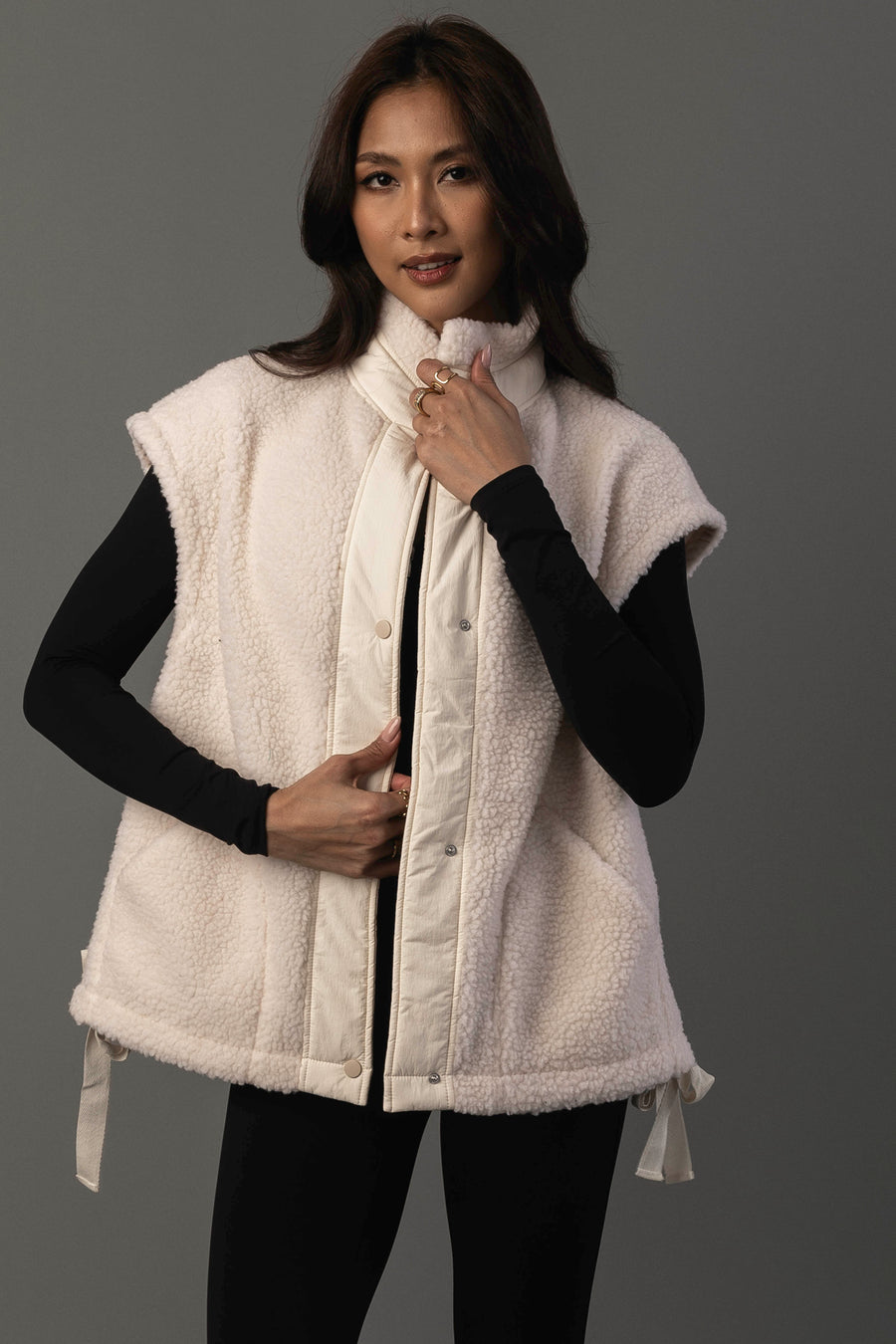 A woman models the Ivory Fleece Vest from Panache Apparel Collection, layering the sleeveless, cream-colored vest over a black long-sleeve shirt and leggings as she poses against a plain gray background, holding the collar with one hand.