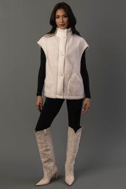 A woman stands against a gray background wearing the Panache Apparel Collection Ivory Fleece Vest over a black long-sleeve top and pants, paired with light-colored knee-high cowboy boots.