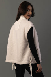 A woman with long dark hair wears the Panache Apparel Collection Ivory Fleece Vest over a black long-sleeve top, posing with her body turned and looking over her shoulder against a gray background.