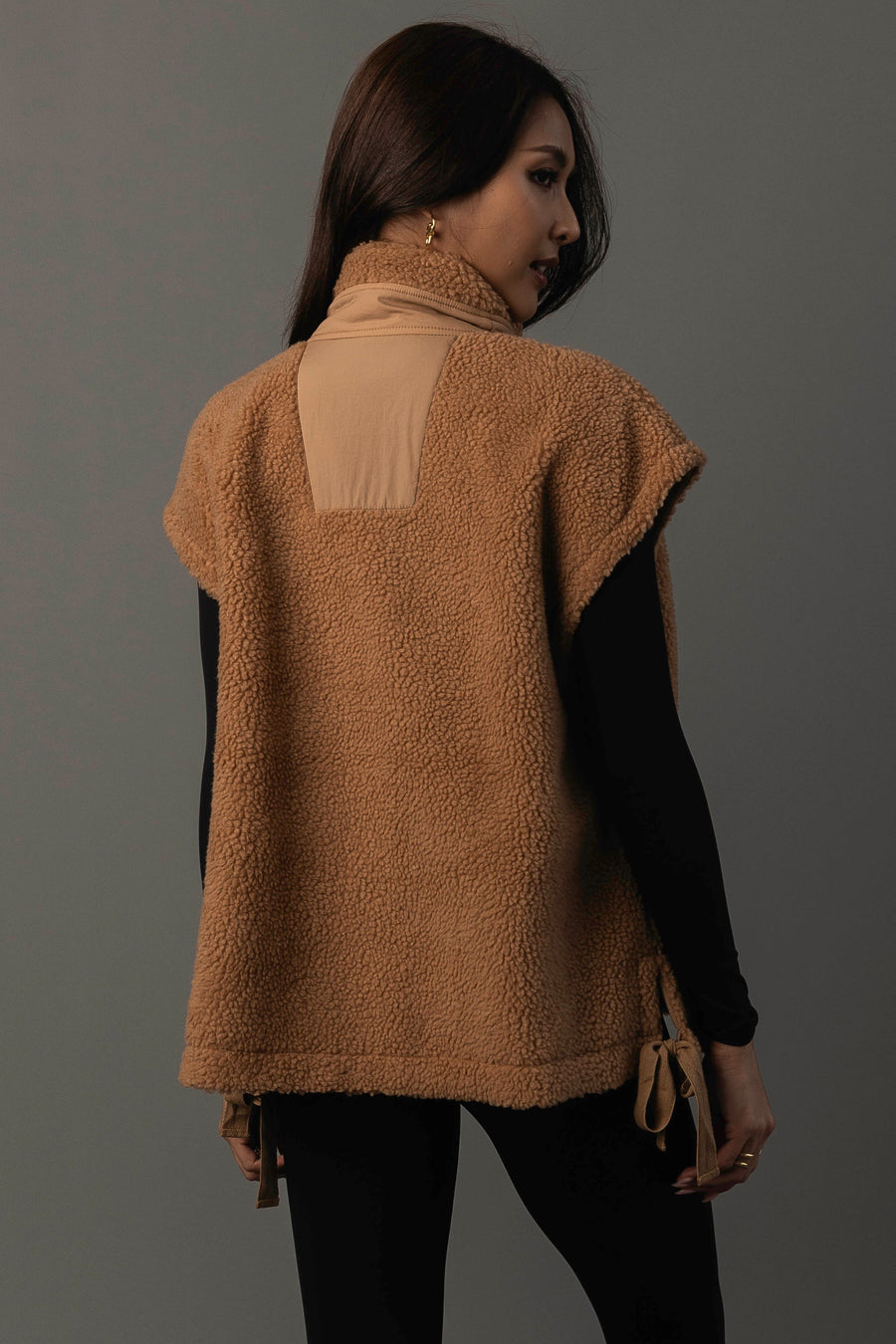 A woman with long dark hair models the Panache Apparel Collection Camel Fleece Vest over a black long-sleeve top, facing away and glancing over her shoulder against a plain gray backdrop.