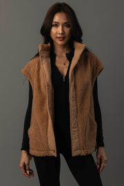 A woman models the Panache Apparel Collection Camel Fleece Vest, a sleeveless brown vest, layered over a black long-sleeve top. She stands against a plain dark gray background, looking at the camera with a slight smile.