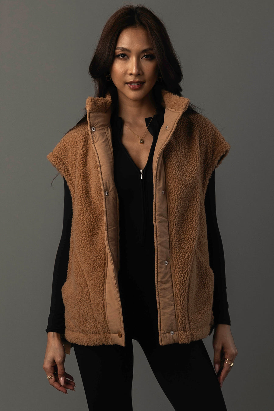 A woman models the Panache Apparel Collection Camel Fleece Vest, a sleeveless brown vest, layered over a black long-sleeve top. She stands against a plain dark gray background, looking at the camera with a slight smile.