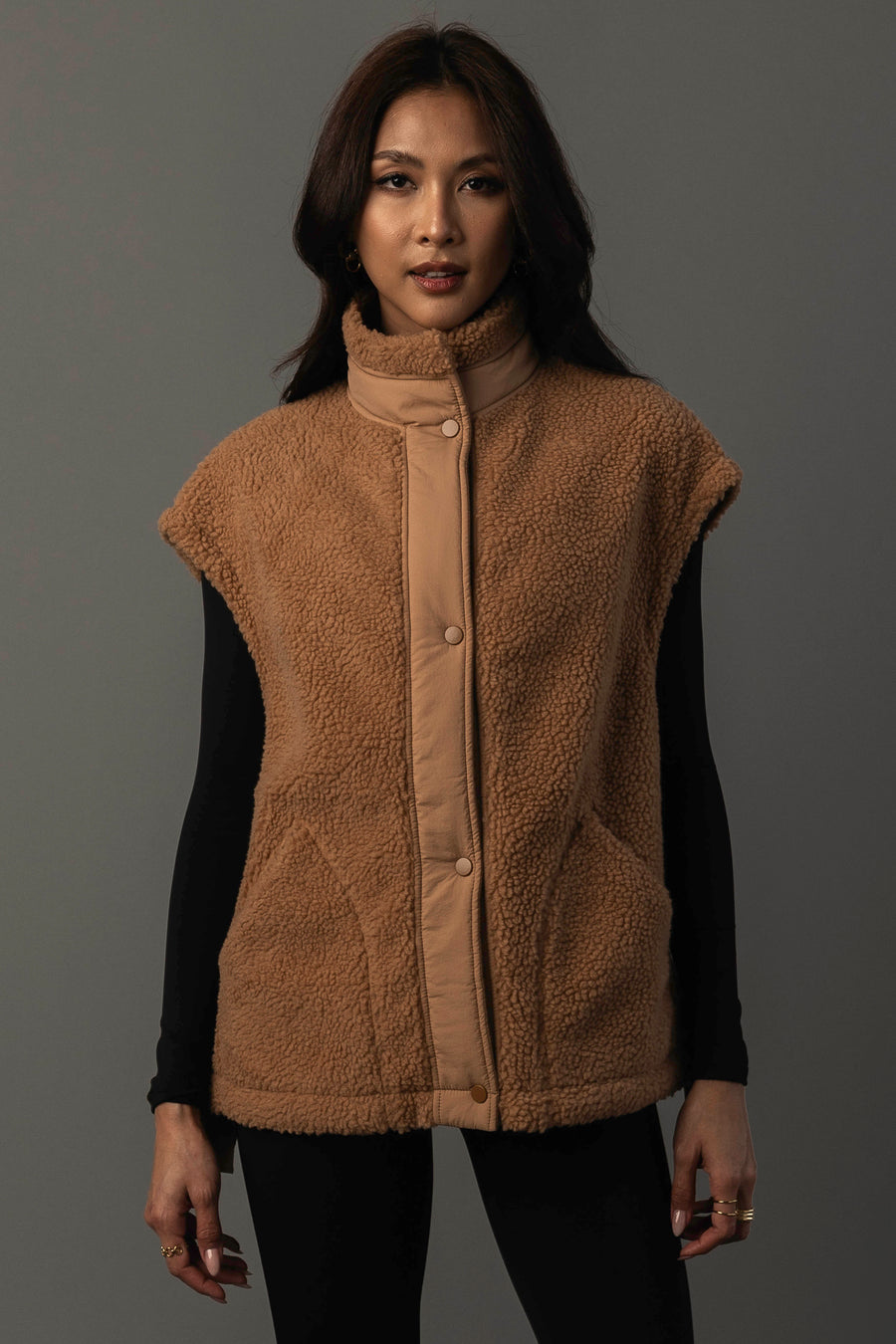 A woman models the Panache Apparel Collection Camel Fleece Vest, layering the tan, sleeveless piece over a black long-sleeve top as she stands against a plain gray background, looking directly at the camera.