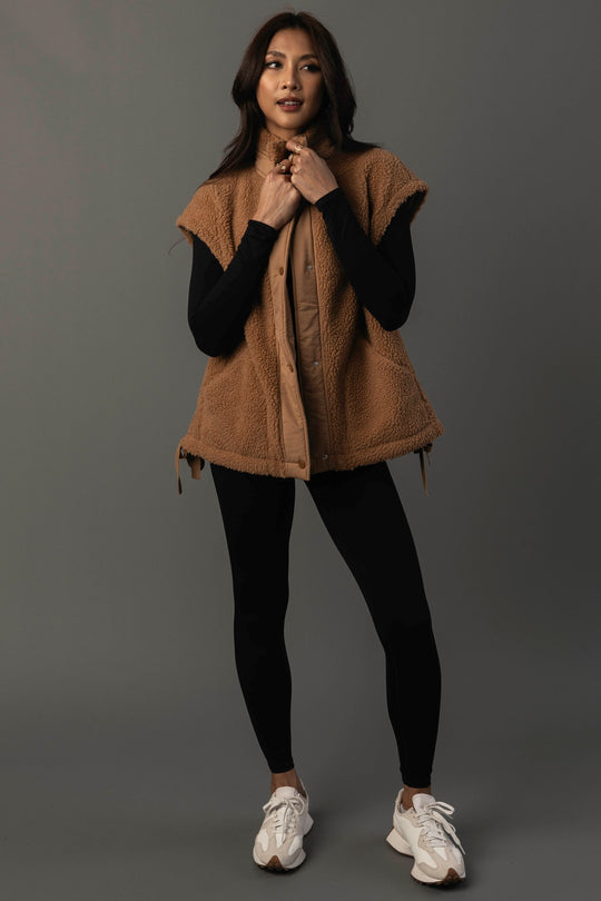 A woman stands against a gray background, wearing the Camel Fleece Vest from Panache Apparel Collection over a black long-sleeve top and black leggings, paired with white sneakers. She looks forward with a slight smile and a relaxed posture.