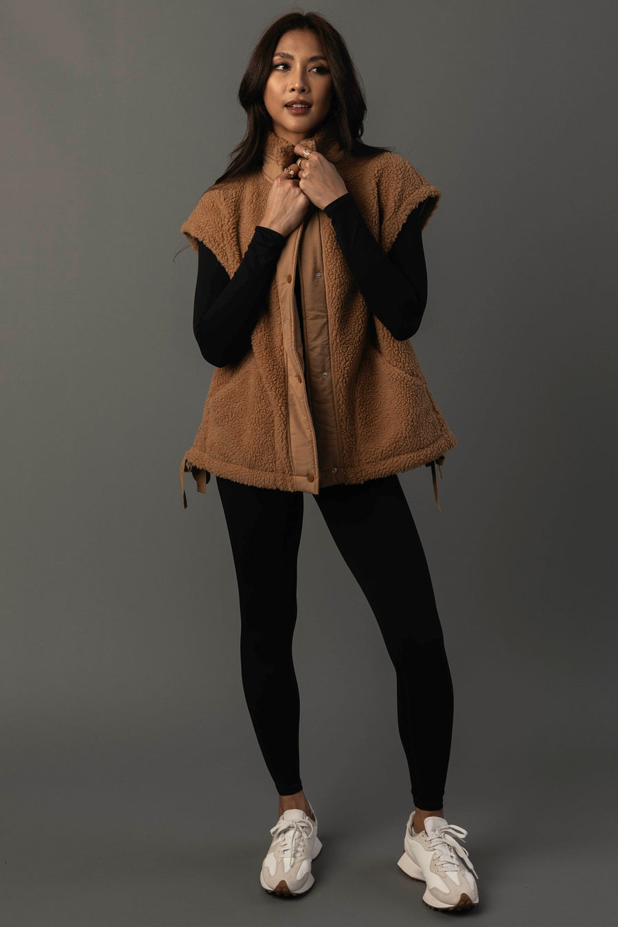 A woman stands against a gray background, wearing the Camel Fleece Vest from Panache Apparel Collection over a black long-sleeve top and black leggings, paired with white sneakers. She looks forward with a slight smile and a relaxed posture.