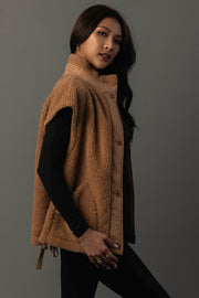A woman with long dark hair stands sideways against a plain gray background, wearing the Panache Apparel Collection Camel Fleece Vest over a black top. She accessorizes with gold hoop earrings and rings, gazing slightly toward the camera.