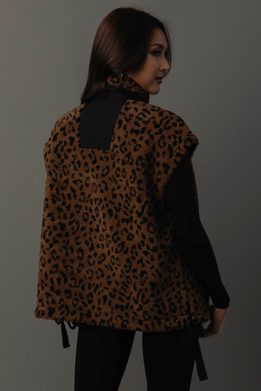 A woman with long dark hair models the Panache Apparel Collection Brown & Black Leopard Fleece Vest over a black outfit, posing with her back turned and glancing over her shoulder against a plain gray background.