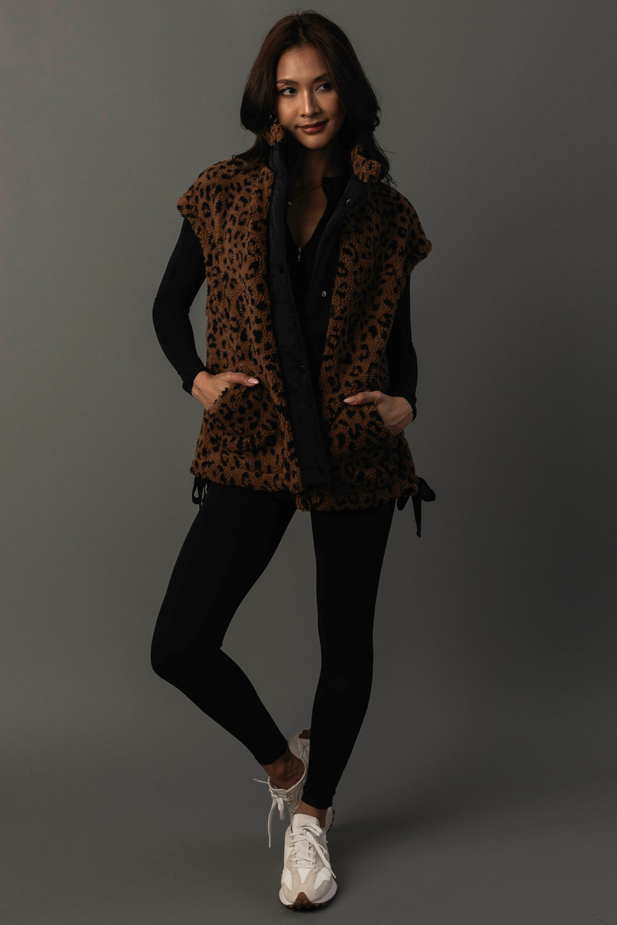 A woman stands against a gray background with her hands in the pockets of the Panache Apparel Collection Brown & Black Leopard Fleece Vest, styled over a black outfit and paired with white sneakers. She smiles gently at the camera.