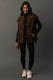 A woman stands and smiles, wearing the Panache Apparel Collection Brown & Black Leopard Fleece Vest over a black long-sleeve top and black leggings, paired with white sneakers, set against a plain dark background.