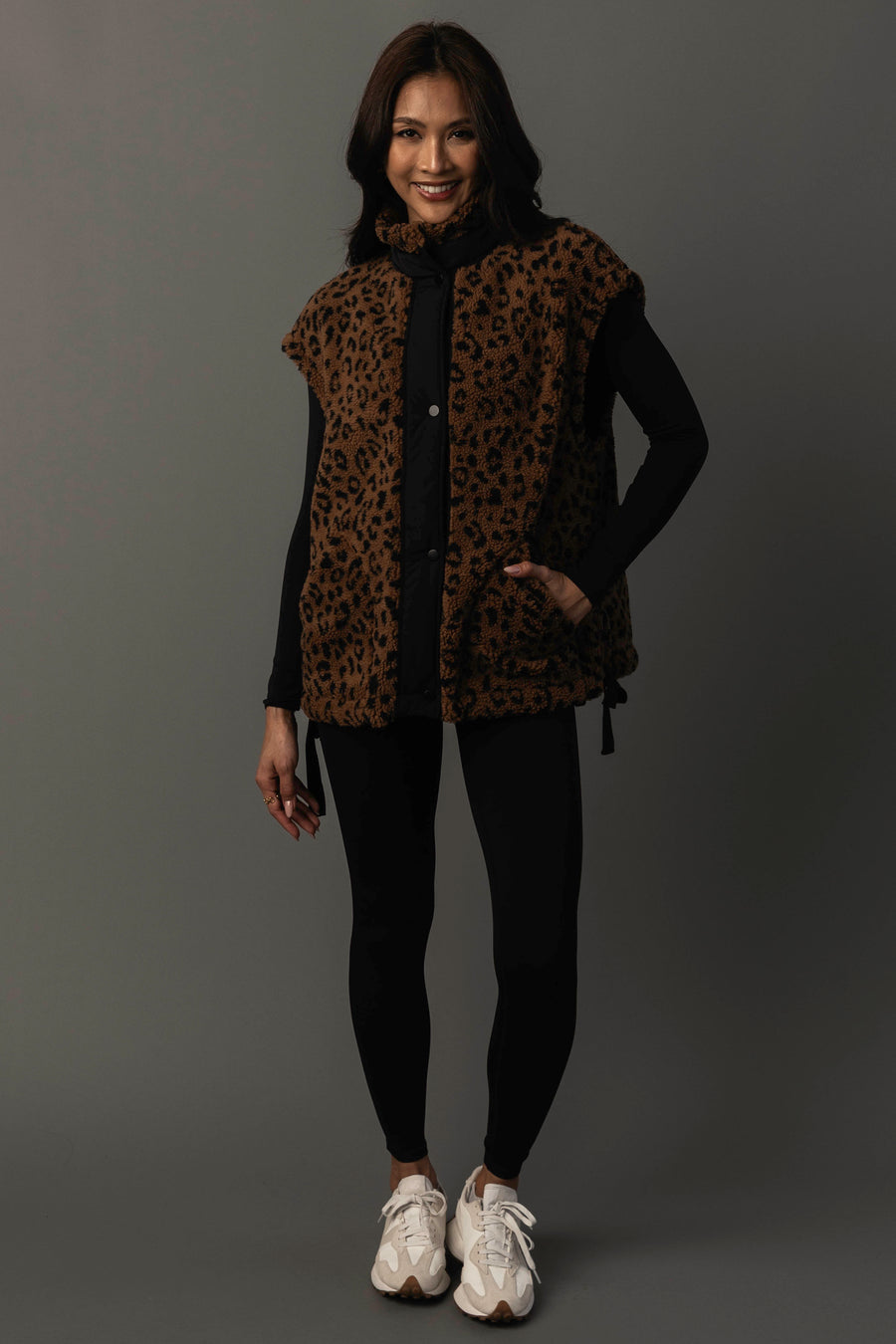 A woman stands and smiles, wearing the Panache Apparel Collection Brown & Black Leopard Fleece Vest over a black long-sleeve top and black leggings, paired with white sneakers, set against a plain dark background.