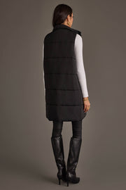 A woman stands facing away against a plain gray background, wearing the Black Long Vest from Panache Apparel Collection over a white long-sleeve top, black leggings, and black knee-high heeled boots.