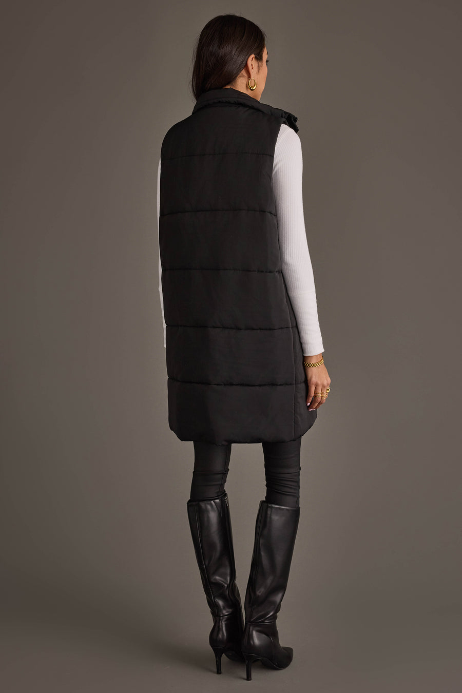 A woman stands facing away against a plain gray background, wearing the Black Long Vest from Panache Apparel Collection over a white long-sleeve top, black leggings, and black knee-high heeled boots.
