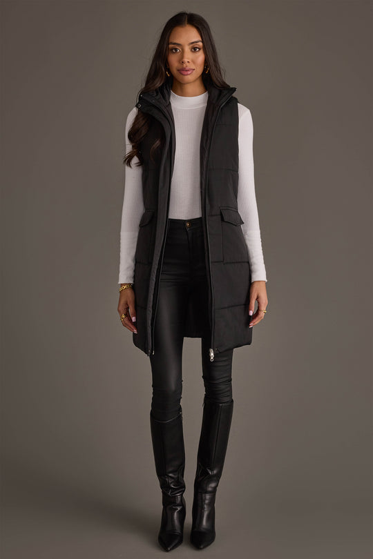 A woman stands facing forward, modeling the Black Long Vest from Panache Apparel Collection over a white turtleneck, paired with black skinny pants and knee-high black boots, against a plain gray background.