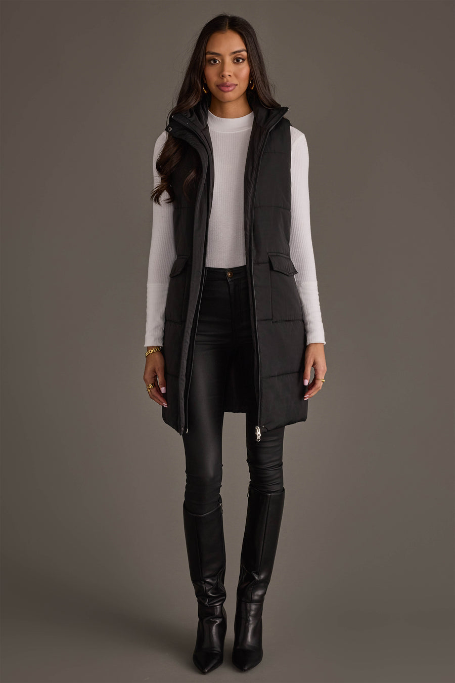 A woman stands facing forward, modeling the Black Long Vest from Panache Apparel Collection over a white turtleneck, paired with black skinny pants and knee-high black boots, against a plain gray background.