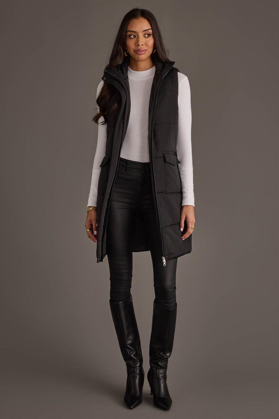 A woman stands facing forward against a plain gray background, wearing the Panache Apparel Collection Black Long Vest over a white ribbed top, black pants, and knee-high black boots.