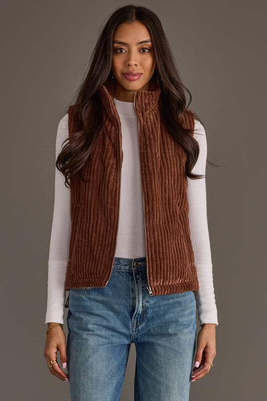 A woman models the Panache Apparel Collection Dark Brown Corduroy Vest, layered over a white long-sleeve shirt and blue jeans. She stands against a gray background, gazing at the camera with her arms relaxed by her sides—an effortless fall layering look.