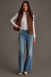 A woman smiles as she poses against a plain gray background, wearing a white long-sleeve top, Panache Apparel Collection’s Dark Brown Corduroy Vest—perfect for fall layering—paired with light blue high-waisted flared jeans and beige heels.