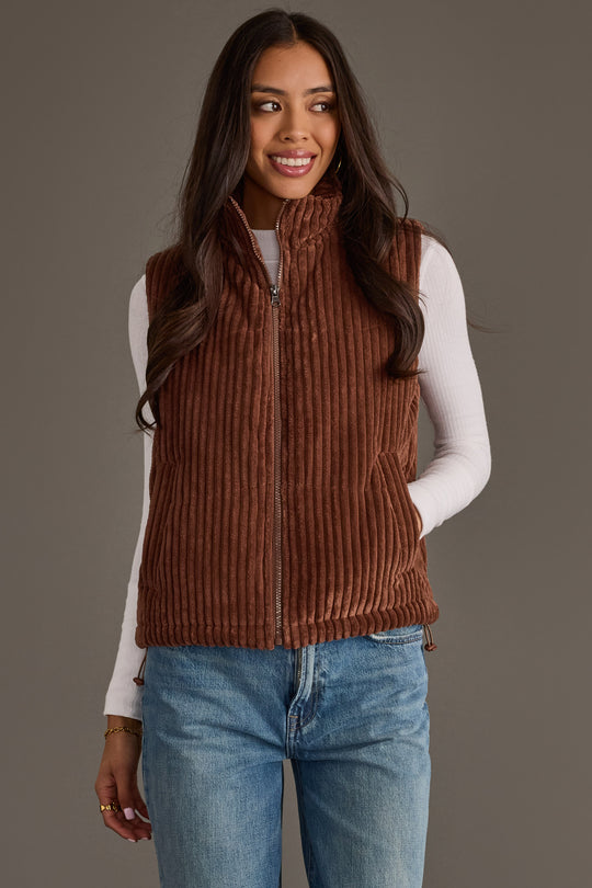 Wearing the Panache Apparel Collection Dark Brown Corduroy Vest over a white long-sleeve shirt and blue jeans, a woman stands against a plain gray background, smiling softly with one hand in her pocket—a relaxed ensemble ideal for fall layering.