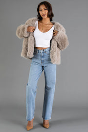 A woman models the Panache Apparel Collection Taupe Faux Fur Jacket, paired with a white crop top, light blue high-waisted jeans, and tan heels against a plain gray background.
