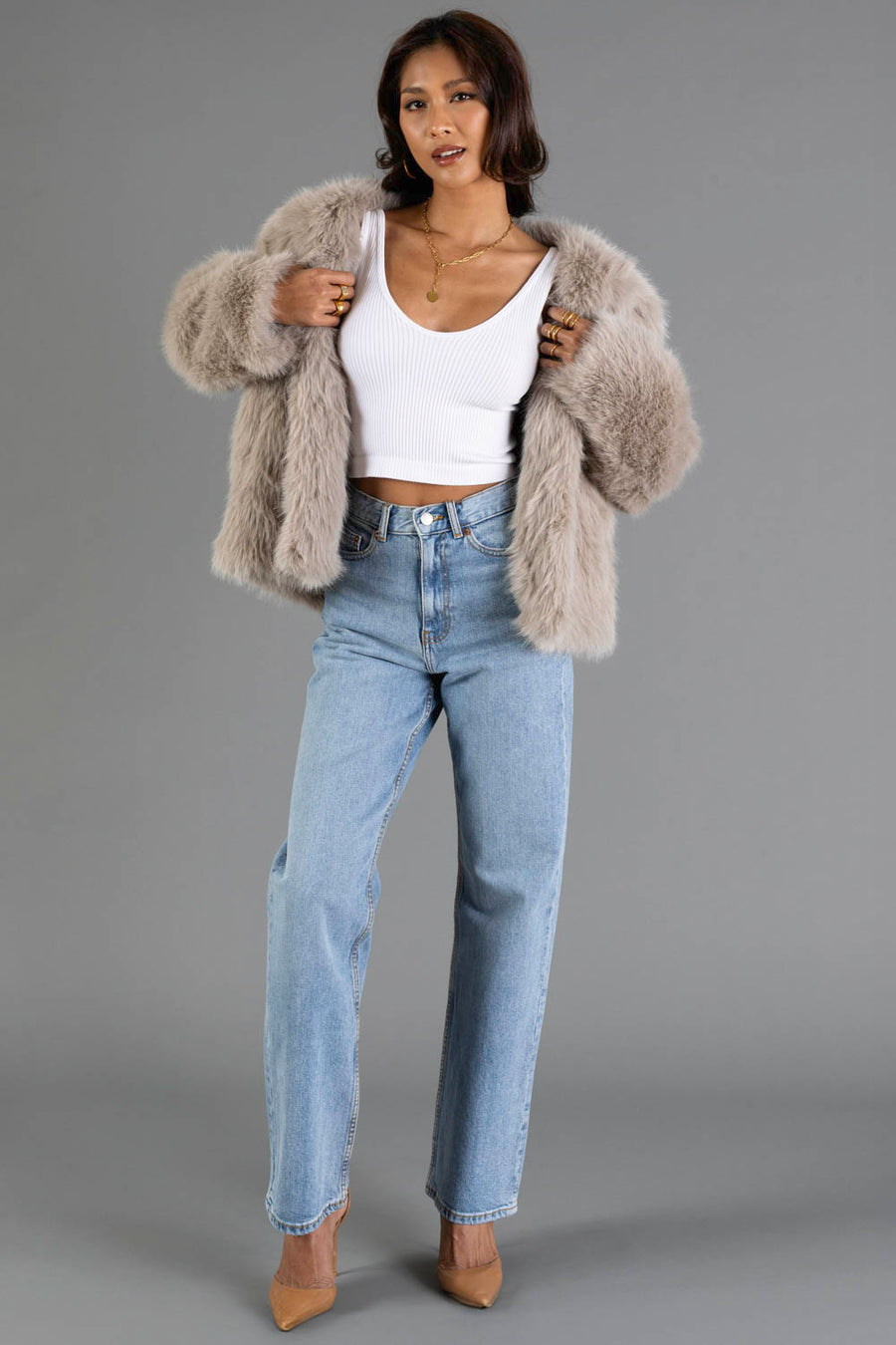 A woman models the Panache Apparel Collection Taupe Faux Fur Jacket, paired with a white crop top, light blue high-waisted jeans, and tan heels against a plain gray background.