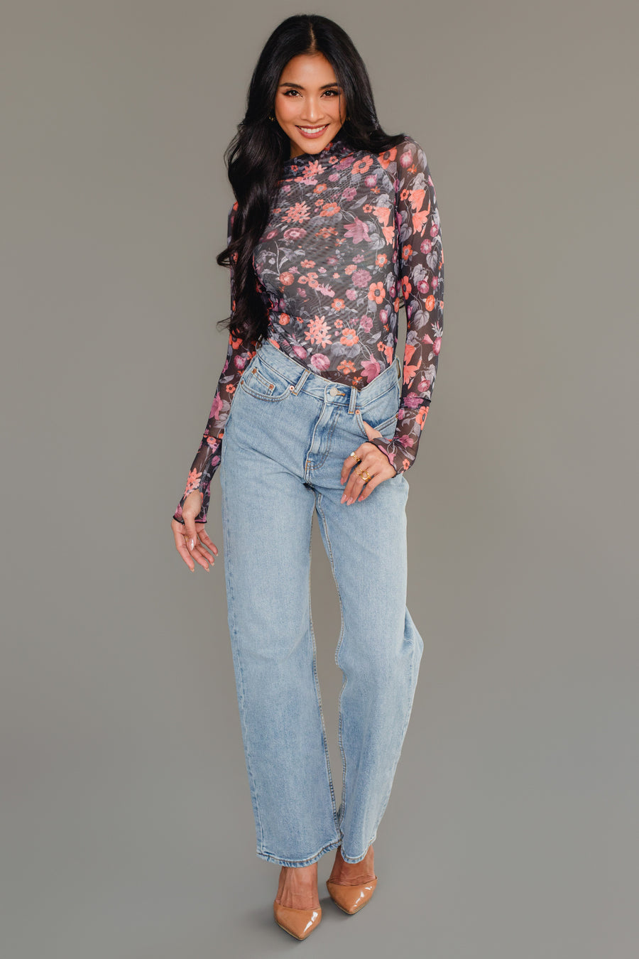 Woman smiling, wearing the Panache Apparel Collection Black, Orange & Pink Floral Mesh Top paired with light blue high-waisted jeans and nude pointed-toe heels, standing against a plain gray background.