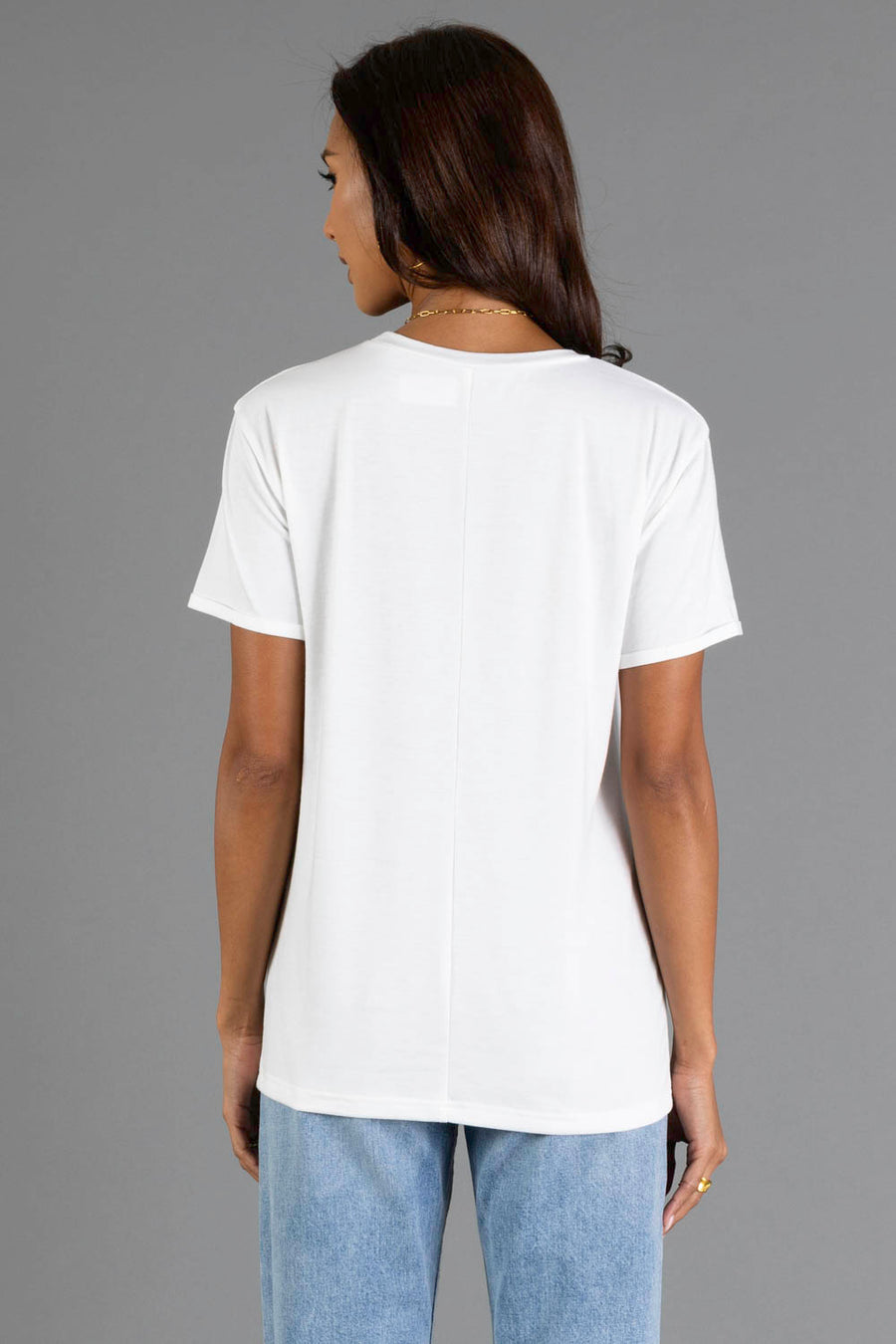 A woman with long dark hair, wearing the Panache Apparel Collection’s Here For The Boos Tee, a plain white short-sleeve t-shirt, and light blue jeans, stands facing away from the camera against a plain gray background.