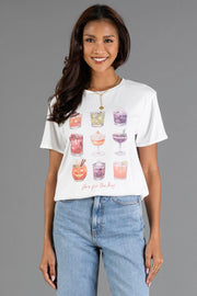 A woman smiles while wearing the Panache Apparel Collection’s “Here For The Boos Tee,” a white t-shirt featuring colorful illustrated cocktails and playful text. She pairs it with blue jeans and stands against a plain gray background.