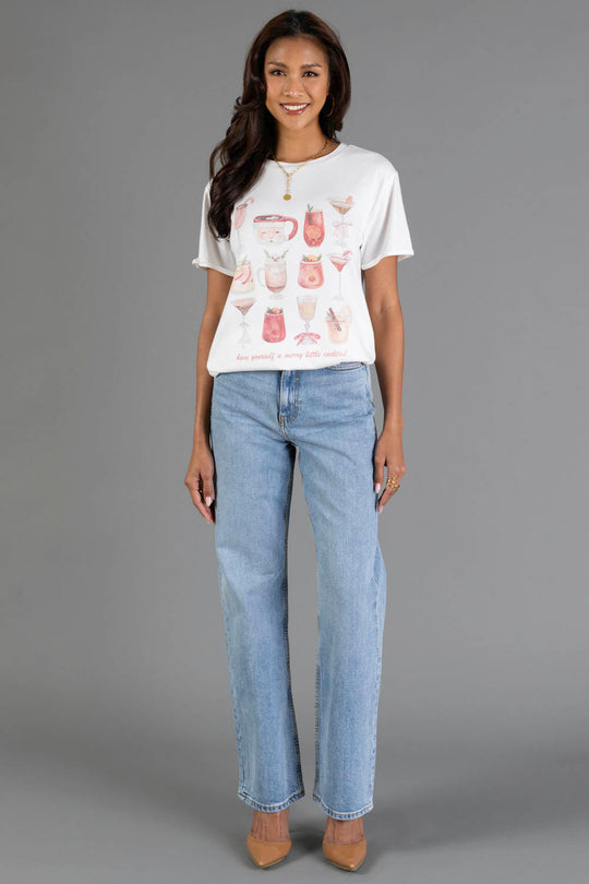 A woman with long, dark hair smiles as she poses against a gray background, wearing the Panache Apparel Collection’s ‘Here For The Boos Tee’, a white printed T-shirt featuring various cocktails, paired with light blue jeans and tan heels.