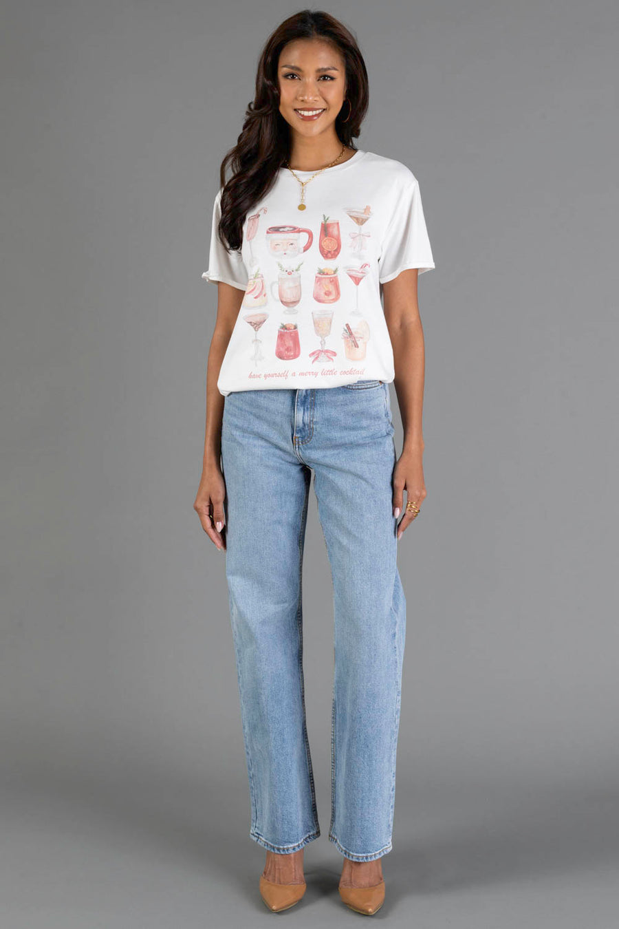 A woman with long, dark hair smiles as she poses against a gray background, wearing the Panache Apparel Collection’s ‘Here For The Boos Tee’, a white printed T-shirt featuring various cocktails, paired with light blue jeans and tan heels.