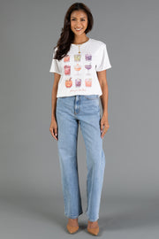 A woman stands and smiles against a plain gray background, wearing the Panache Apparel Collection’s “Here For The Boos Tee”—a white graphic t-shirt featuring colorful drinks—paired with light blue jeans and beige heels.