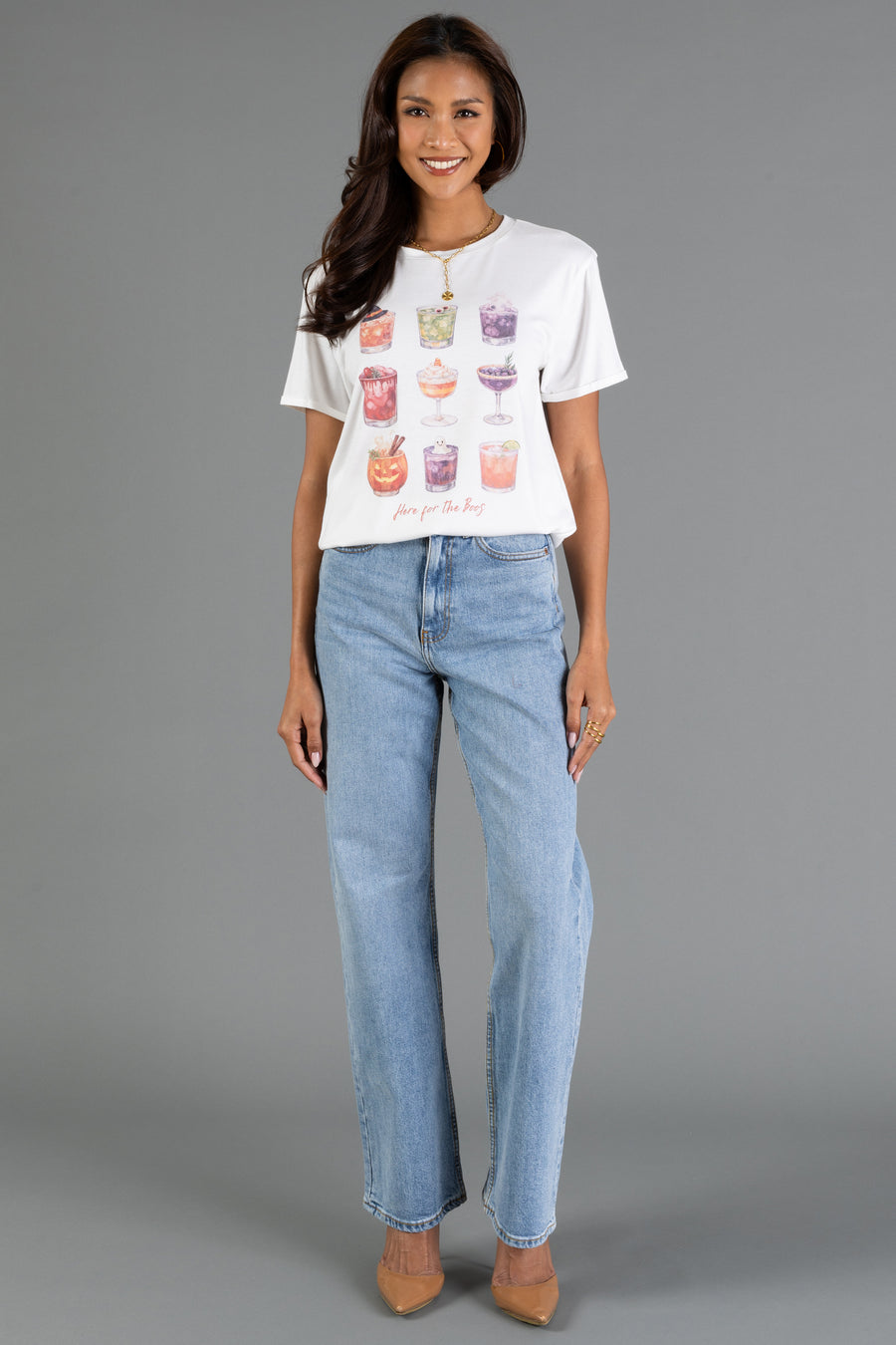 A woman stands and smiles against a plain gray background, wearing the Panache Apparel Collection’s “Here For The Boos Tee”—a white graphic t-shirt featuring colorful drinks—paired with light blue jeans and beige heels.