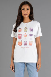 A woman wearing the Panache Apparel Collection “Here For The Boos Tee,” featuring watercolor illustrations of colorful drinks and playful text, styled with light blue jeans and gold jewelry, poses against a gray background.