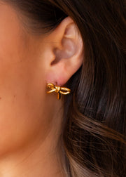 Close-up of a woman's ear wearing panacheaccessories' Luxe Gold Bow Stud Earrings, partially concealed by her brown hair. These hypoallergenic studs offer everyday elegance and comfort.